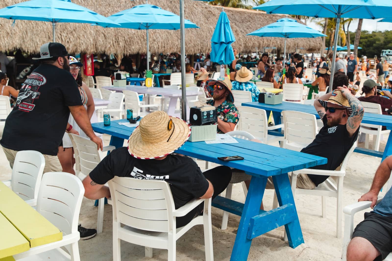 People in straw hats and Bonanza shirts talking at blue picnic tables under tiki umbrellas