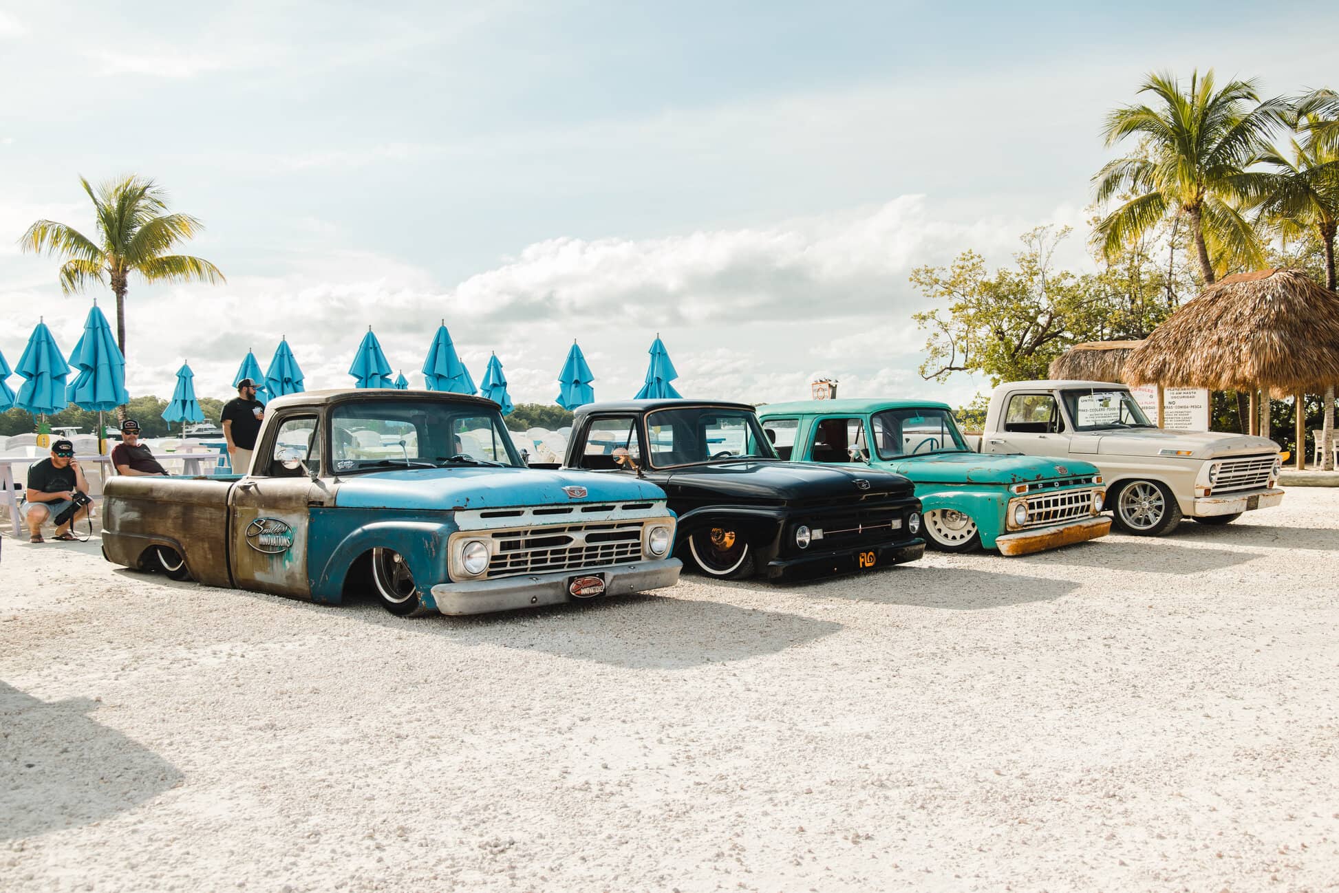 Classic trucks lined up on white sand beside turquoise umbrellas